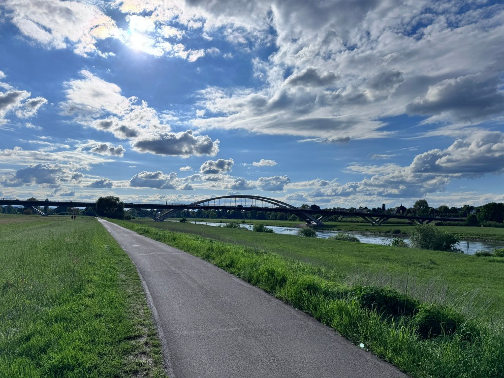 A scenic view of a pathway alongside a river, with a bridge in the background under a blue sky filled with fluffy clouds.