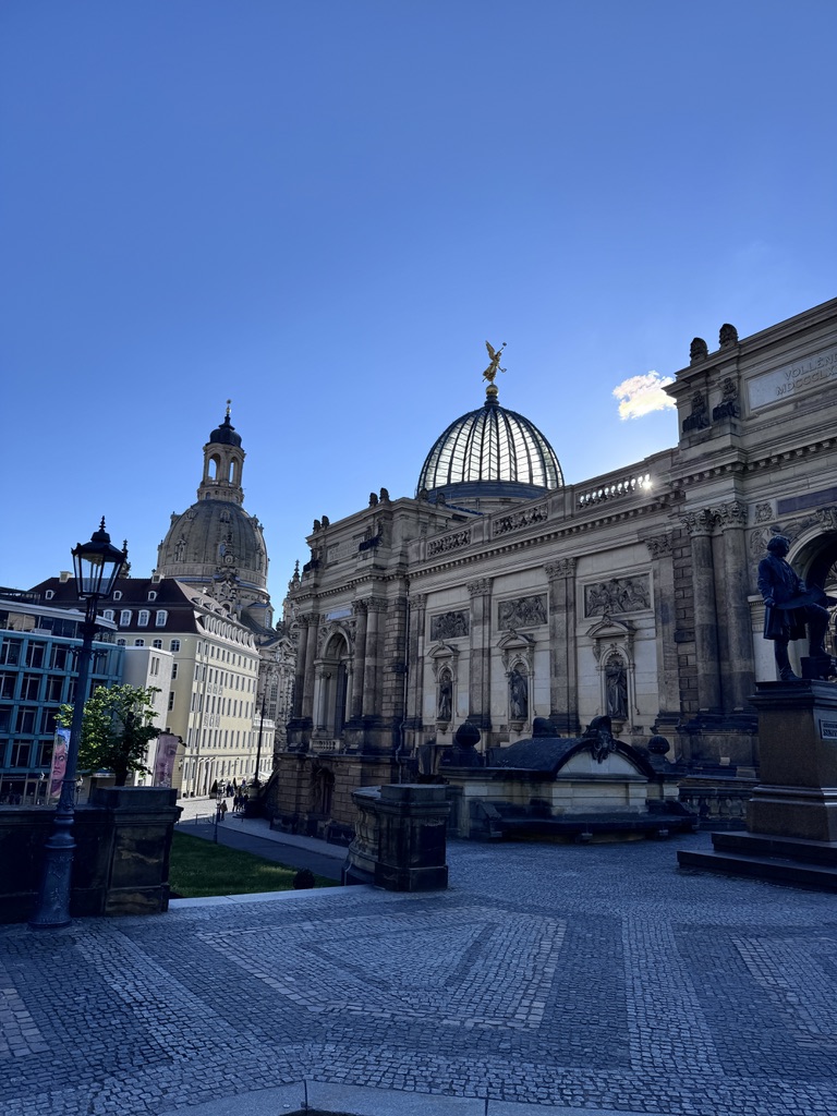 Historic architecture featuring a grand dome with a golden statue, surrounded by cobblestone streets and modern buildings under a clear blue sky.