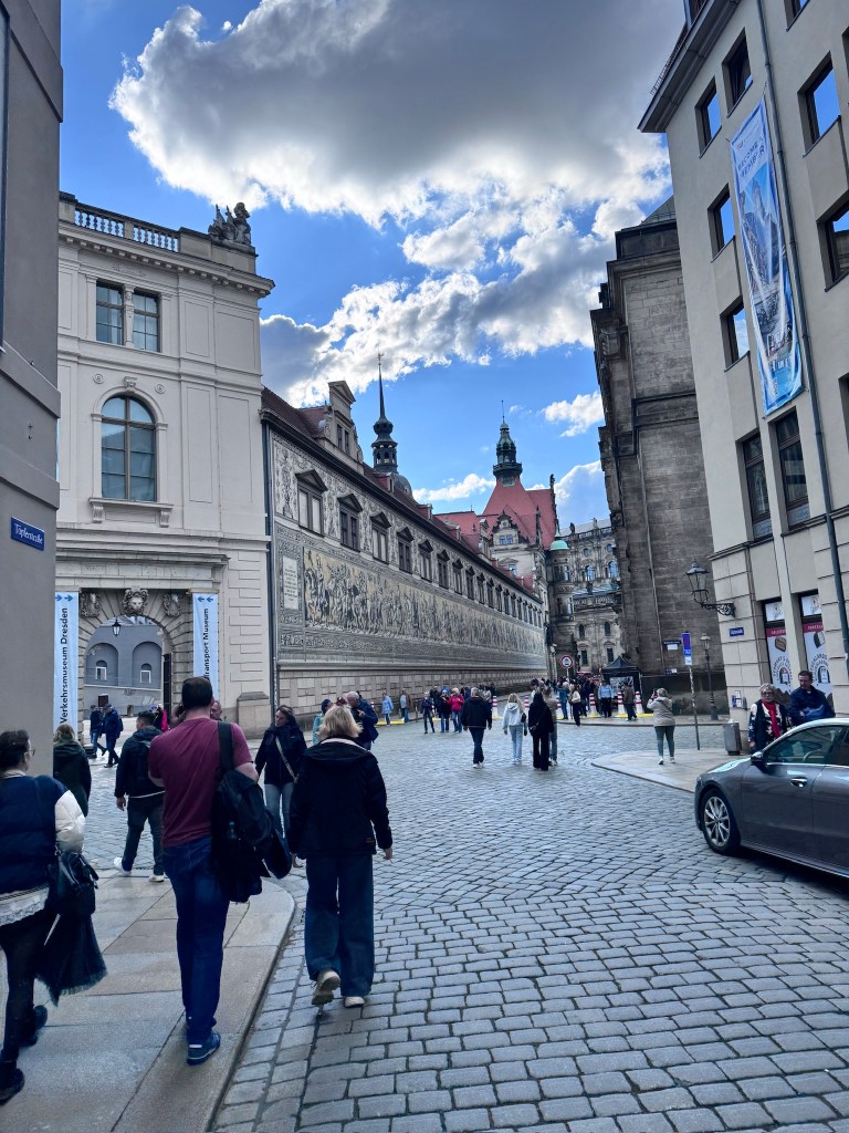A cobblestone street lined with historic buildings and a blue sky with clouds, featuring pedestrians walking and a parked car.