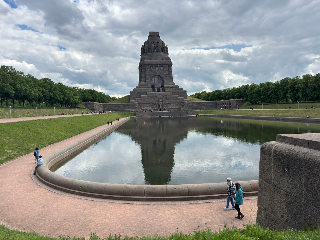 A large monument reflected in a calm pond, surrounded by people walking on a path, with green trees and a cloudy sky in the background.