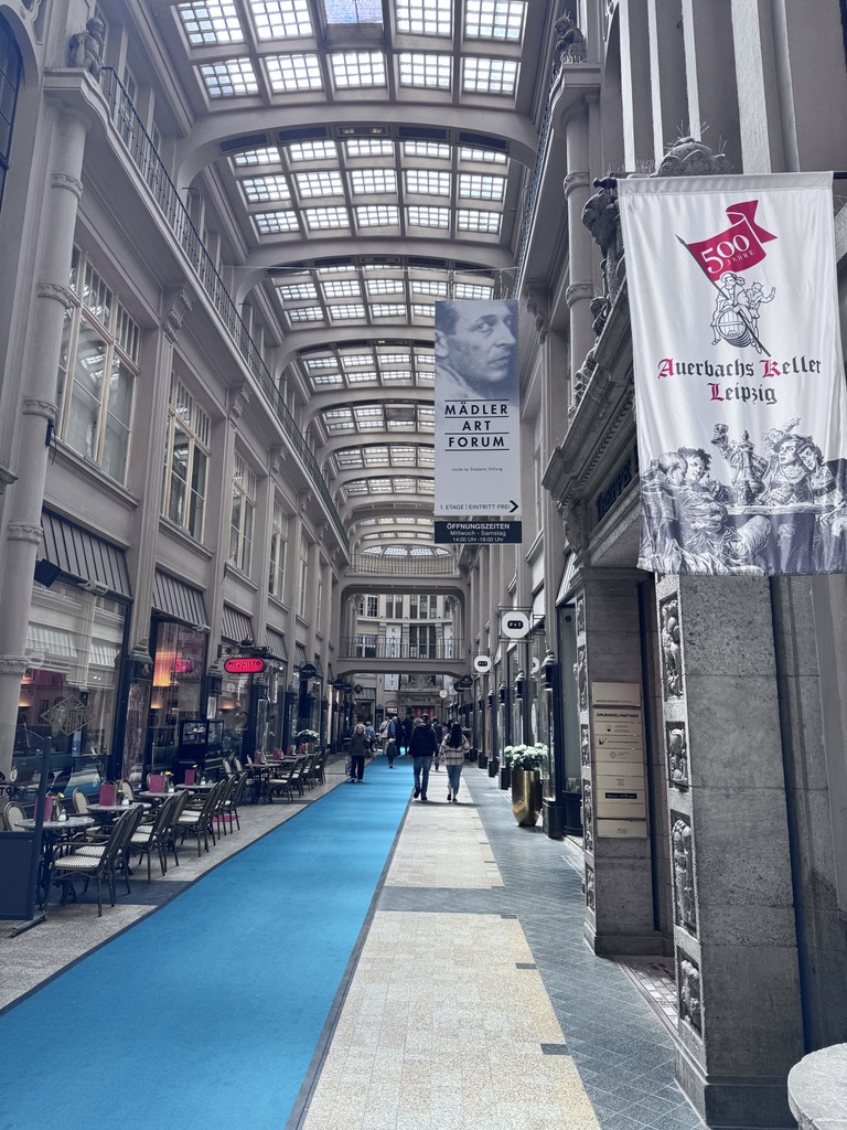 A wide hallway with a blue carpet, lined with tables and chairs on one side. Banners for Mädler Art Forum and Auerbachs Keller are visible, along with large windows letting in natural light from above.