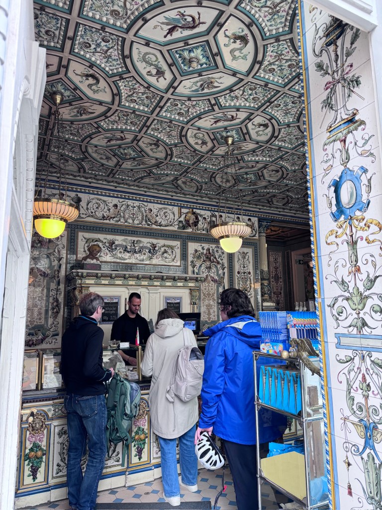 Interior view of a shop featuring a beautifully decorated ceiling and walls, with detailed patterns and colors. Two customers are standing at the counter, interacting with a staff member.