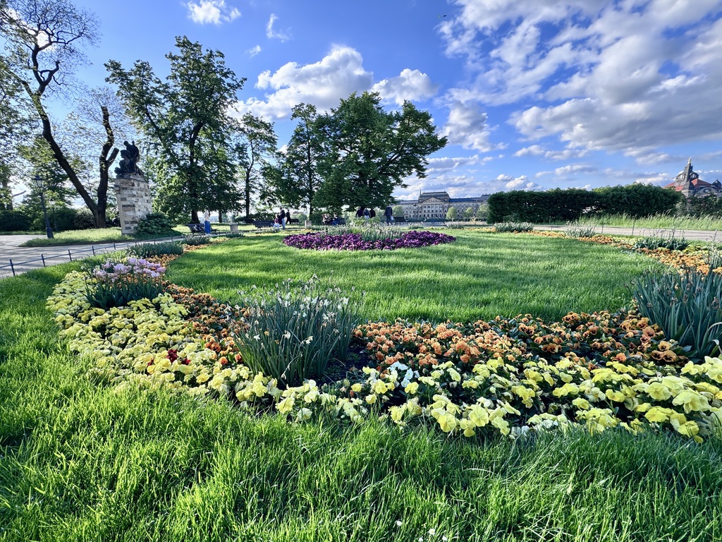 A landscaped garden featuring vibrant flowerbeds arranged in circular patterns, surrounded by lush green grass and trees, under a blue sky with scattered clouds.
