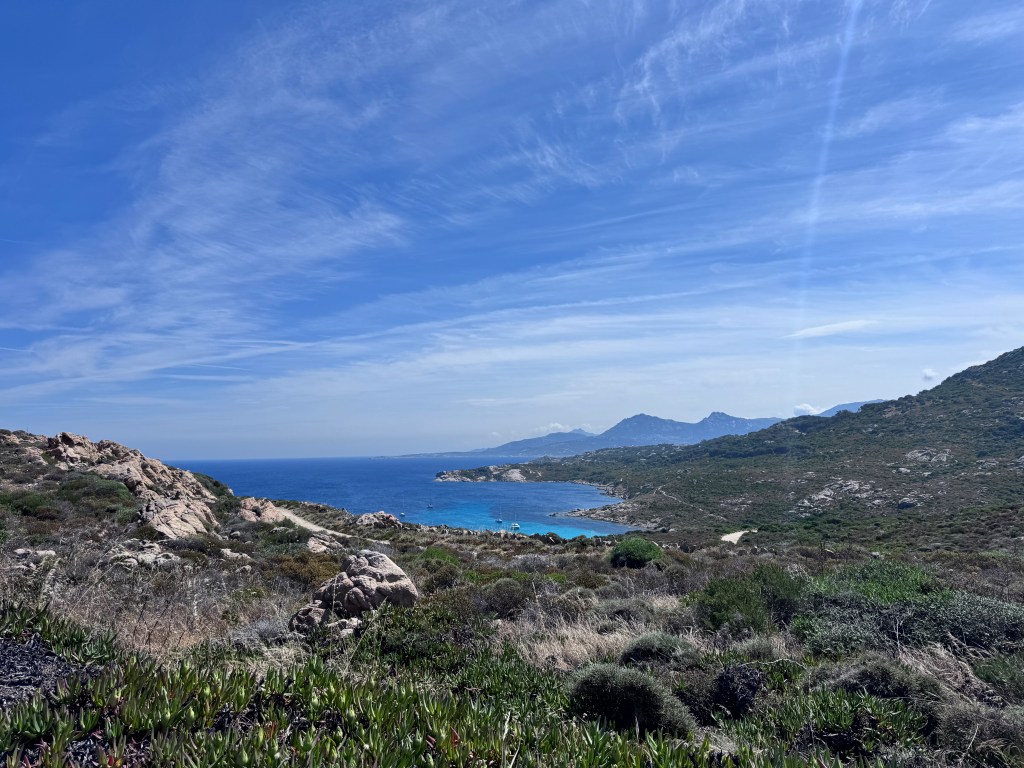 Scenic coastal landscape featuring rocky cliffs, turquoise water, and lush greenery under a blue sky with scattered clouds.