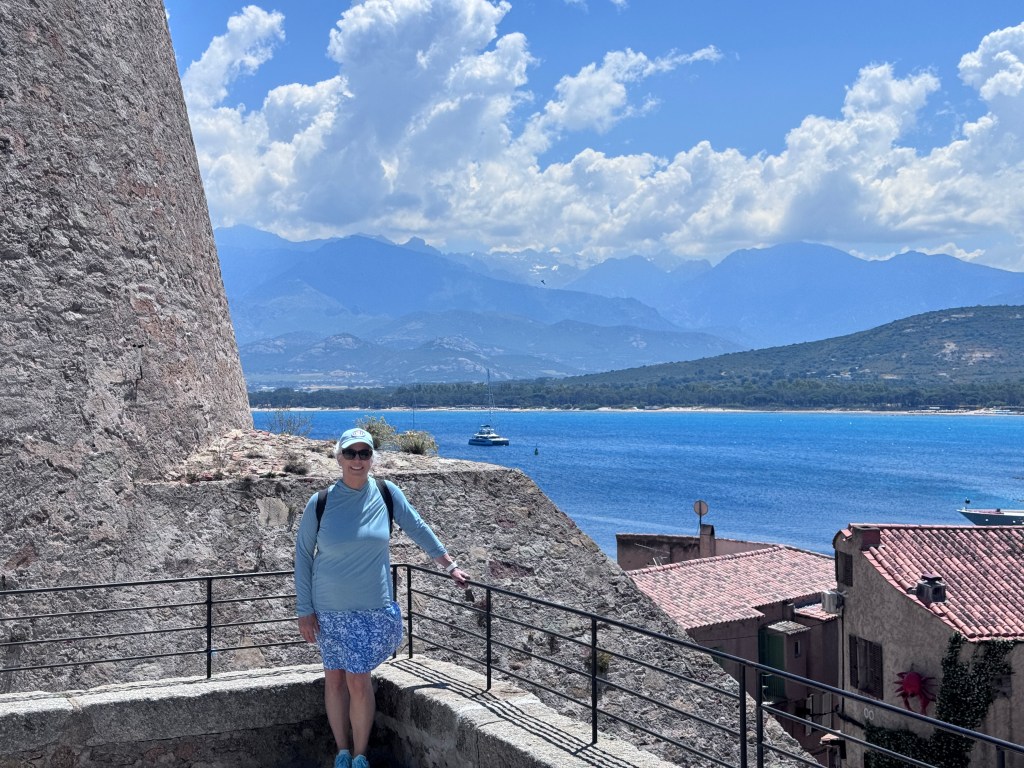 A person stands near a stone structure with a railing, overlooking a blue sea and mountains in the background, under a partly cloudy sky.