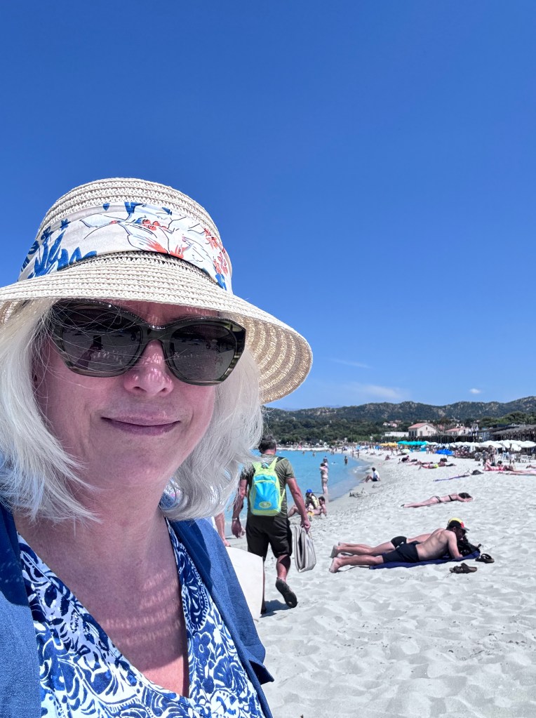 A woman with blonde hair wearing a straw hat and sunglasses smiles at the beach, with people enjoying the sun and swimming in the background.