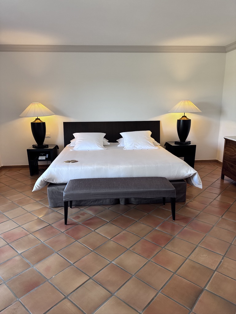 A modern hotel bedroom featuring a large bed with white linens, two black bedside lamps, and a gray bench at the foot of the bed, set against a tiled floor.