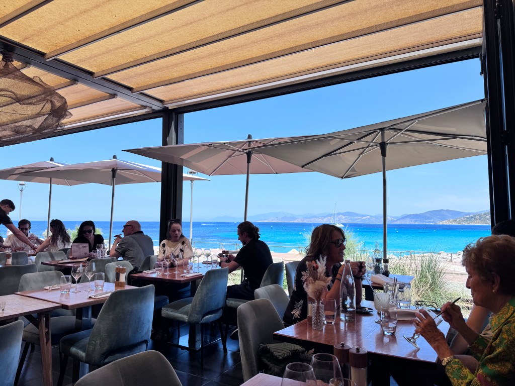 Interior view of a beachside restaurant with patrons dining at tables, large umbrellas providing shade, and a panoramic ocean view in the background.
