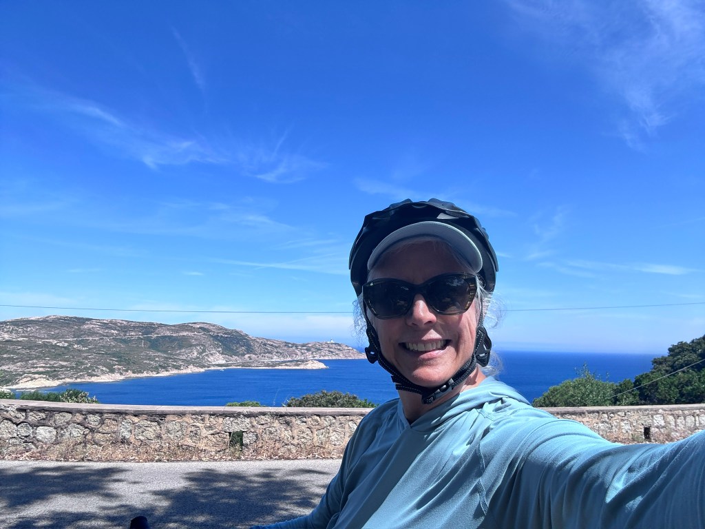 A person wearing a helmet and sunglasses takes a selfie by the coast, with a blue sea and sky visible in the background.