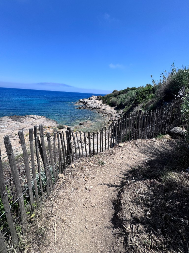 A coastal path lined with a wooden fence along rocky shores and clear blue waters under a bright sky.