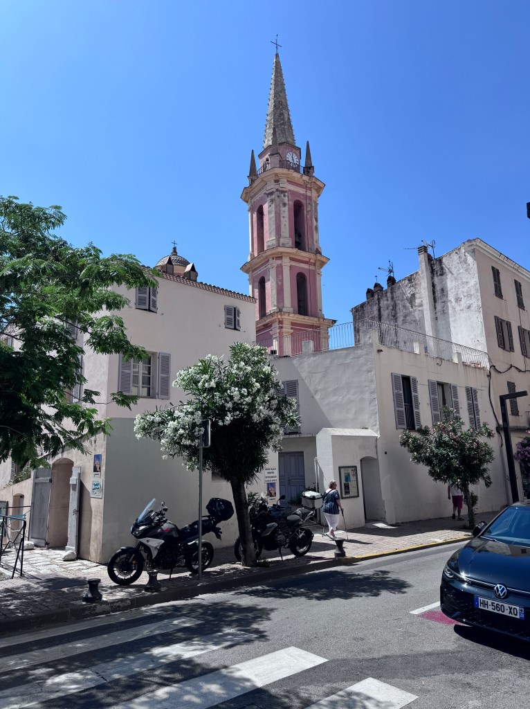 A view of a charming street featuring a tall pink church tower with a spire against a clear blue sky, framed by greenery and parked motorcycles.