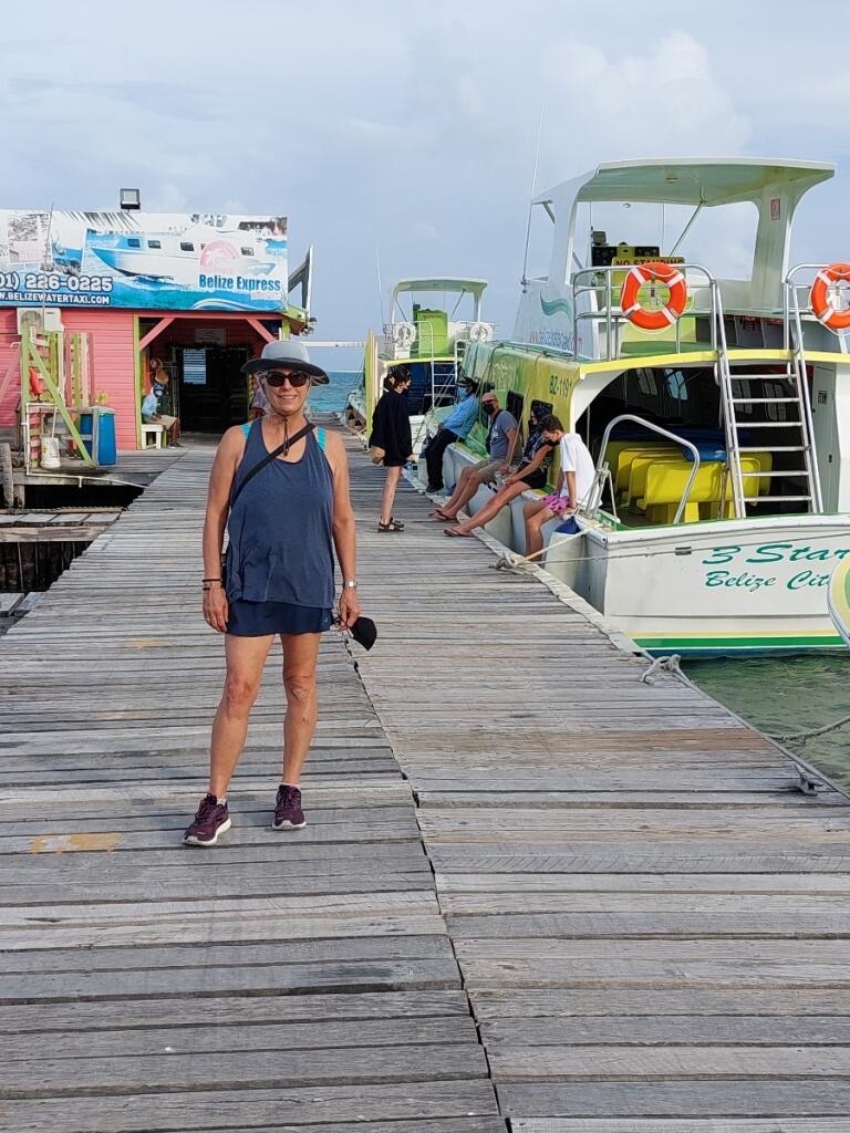 A woman stands on a wooden pier beside colorful boats with a waterside building in the background, under a cloudy sky.