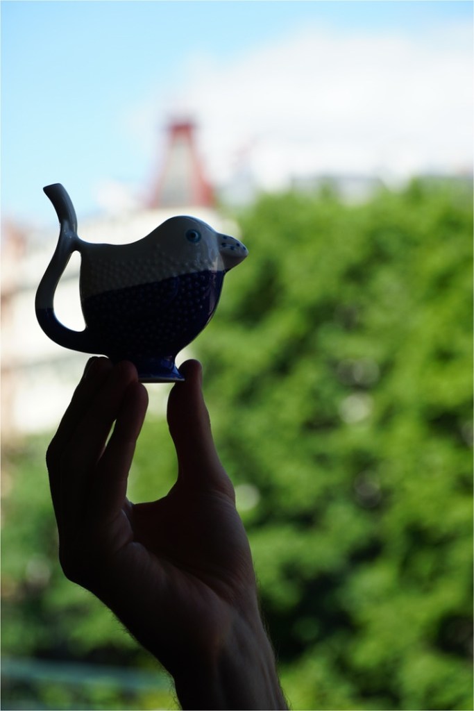A hand holding a decorative ceramic bird-shaped teapot against a blurred green background.