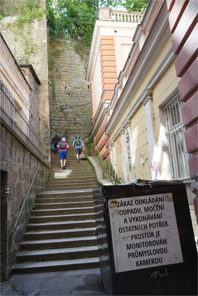 A view of a steep staircase surrounded by old buildings, with three people walking up the stairs. A trash bin is visible in the foreground, displaying a sign in Czech that warns against littering and indicates surveillance.