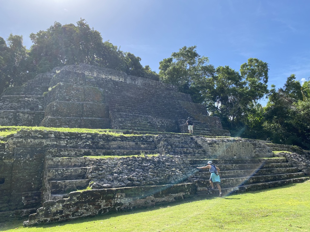 Two people exploring an ancient stone pyramid surrounded by lush greenery under a bright blue sky.