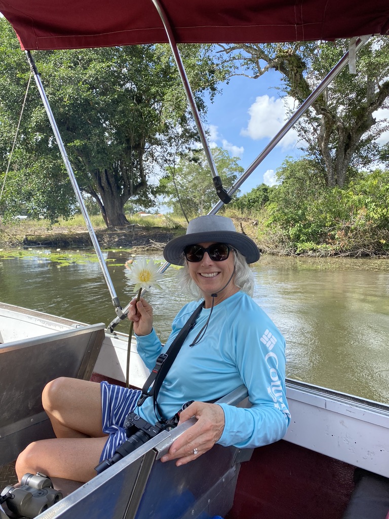 Smiling woman sitting on a boat, wearing a sun hat and long-sleeve shirt, holding a flower with a camera beside her.