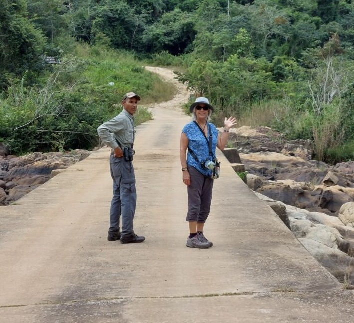 Two people standing on a stone bridge in a lush, green landscape. One person is waving and smiling, while the other looks back towards the camera.