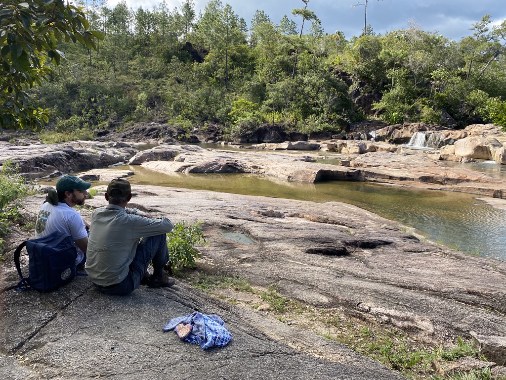 Two people sitting on a rocky riverbank, overlooking a calm water body surrounded by greenery and trees.