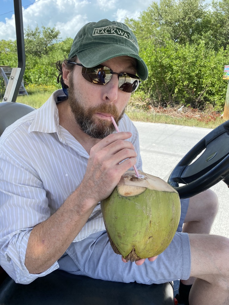 A man sitting in a golf cart, sipping from a coconut with a straw, wearing sunglasses and a green cap, dressed in a striped shirt and shorts, with greenery in the background.