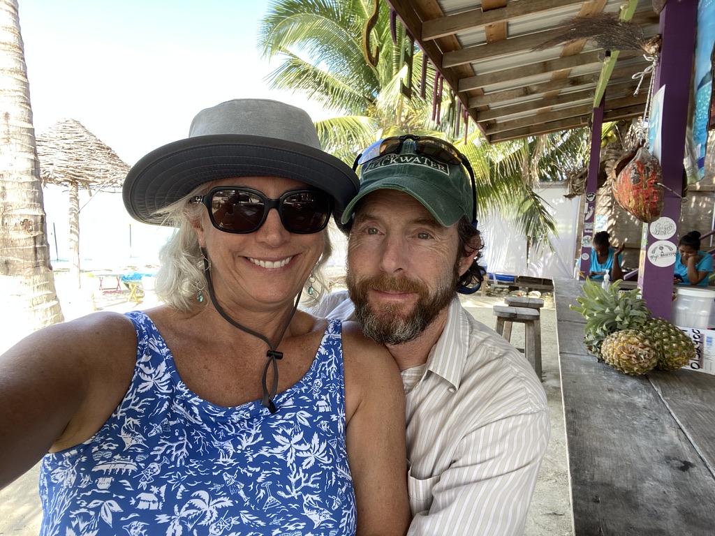 A smiling couple posing for a selfie in a tropical setting, with palm trees and a beach in the background.