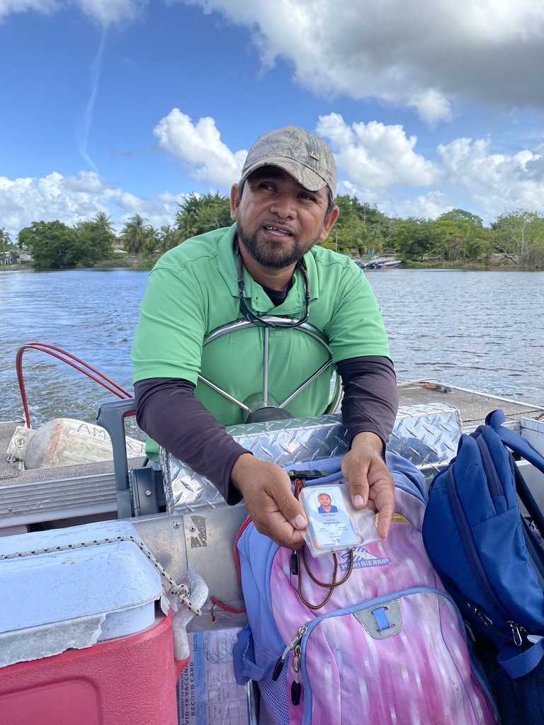 A man in a green shirt holds up an identification card while sitting in a boat, surrounded by backpacks and the river in the background under partly cloudy skies.