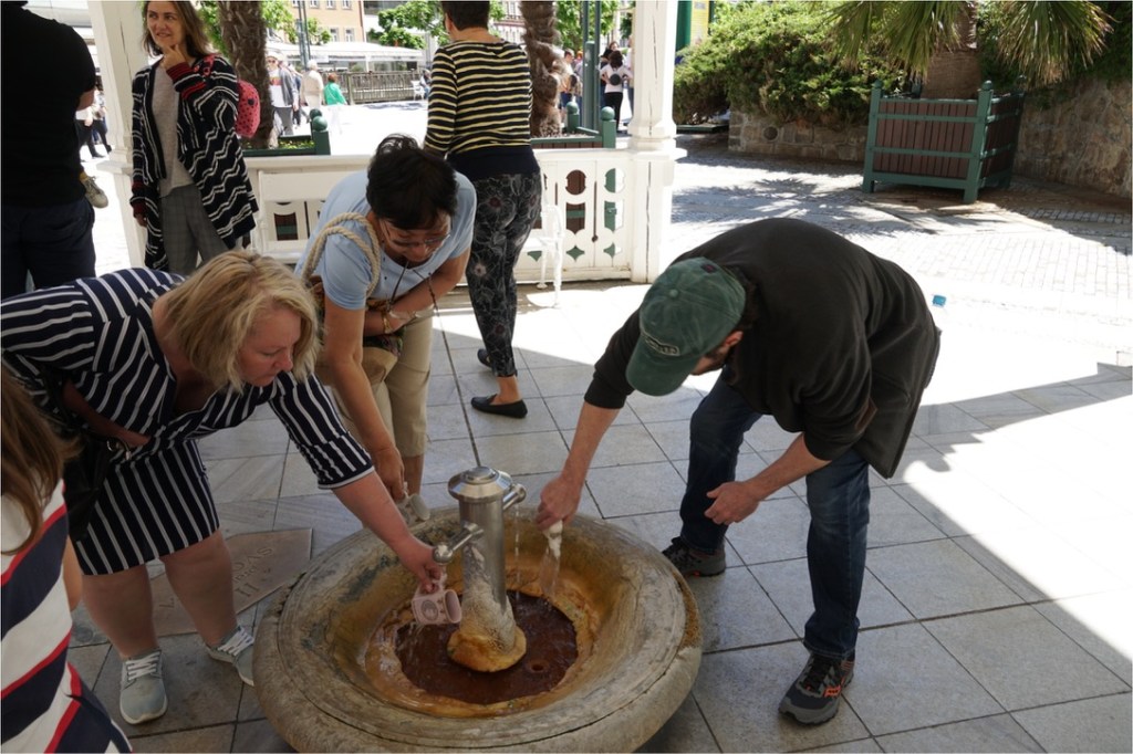 A group of people engaging with a traditional water fountain, with one person operating the fountain while others observe and assist.