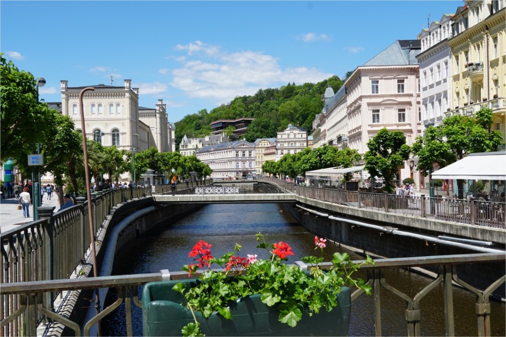 A scenic view of a canal lined with trees and vibrant buildings, with people walking along the promenade under a clear blue sky.