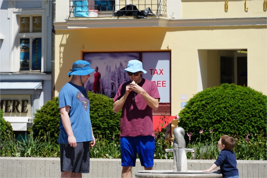 Two men wearing blue hats interact near a water fountain, while a small child observes from the side.