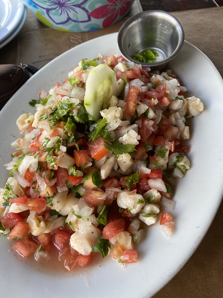 A plate of fresh ceviche topped with diced tomatoes, onions, cilantro, and cucumber, served with a side of lime in a small metal container.